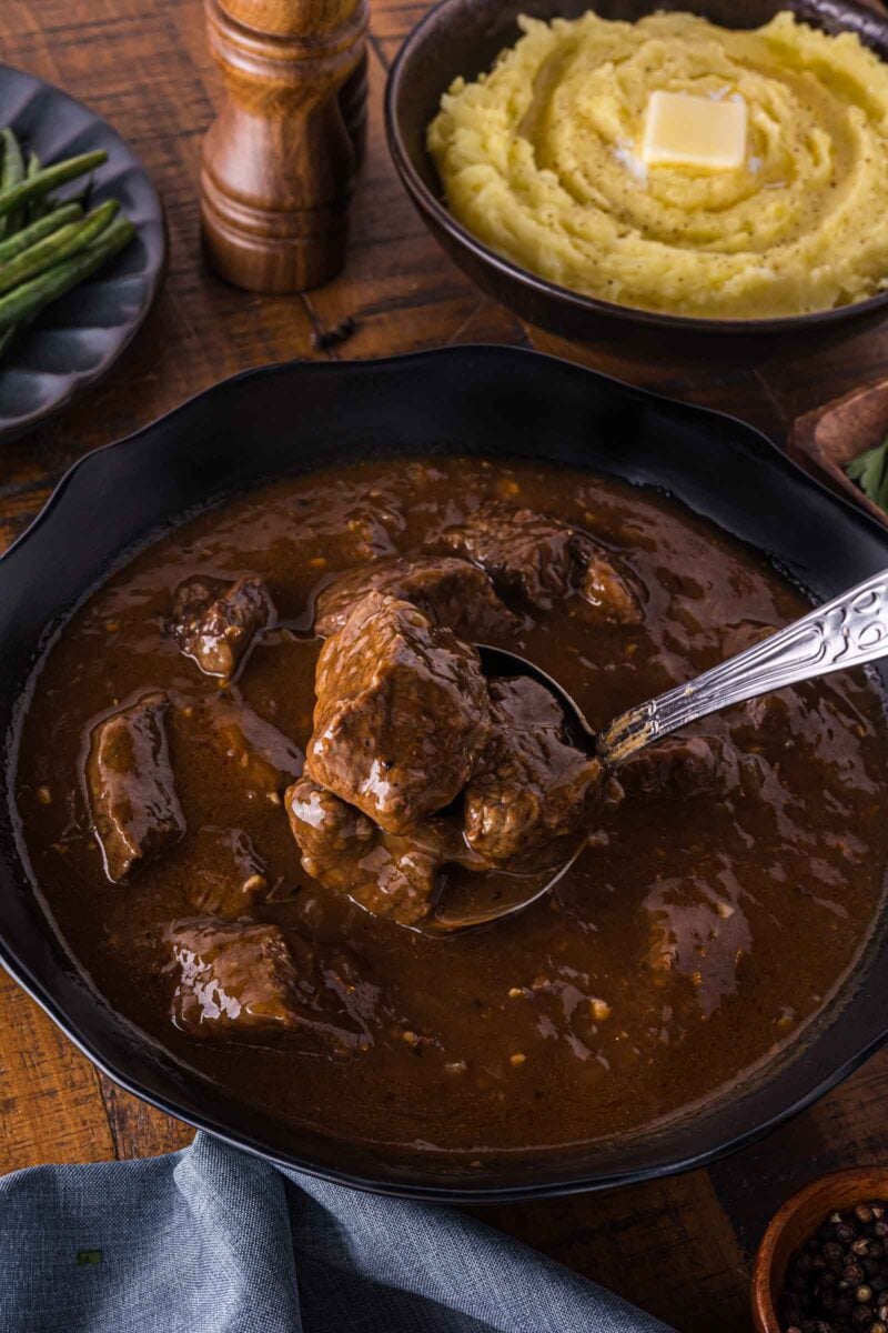 Bowl of beef tips in brown gravy, with a spoon lifting a portion; mashed potatoes with butter and green beans are in the background.