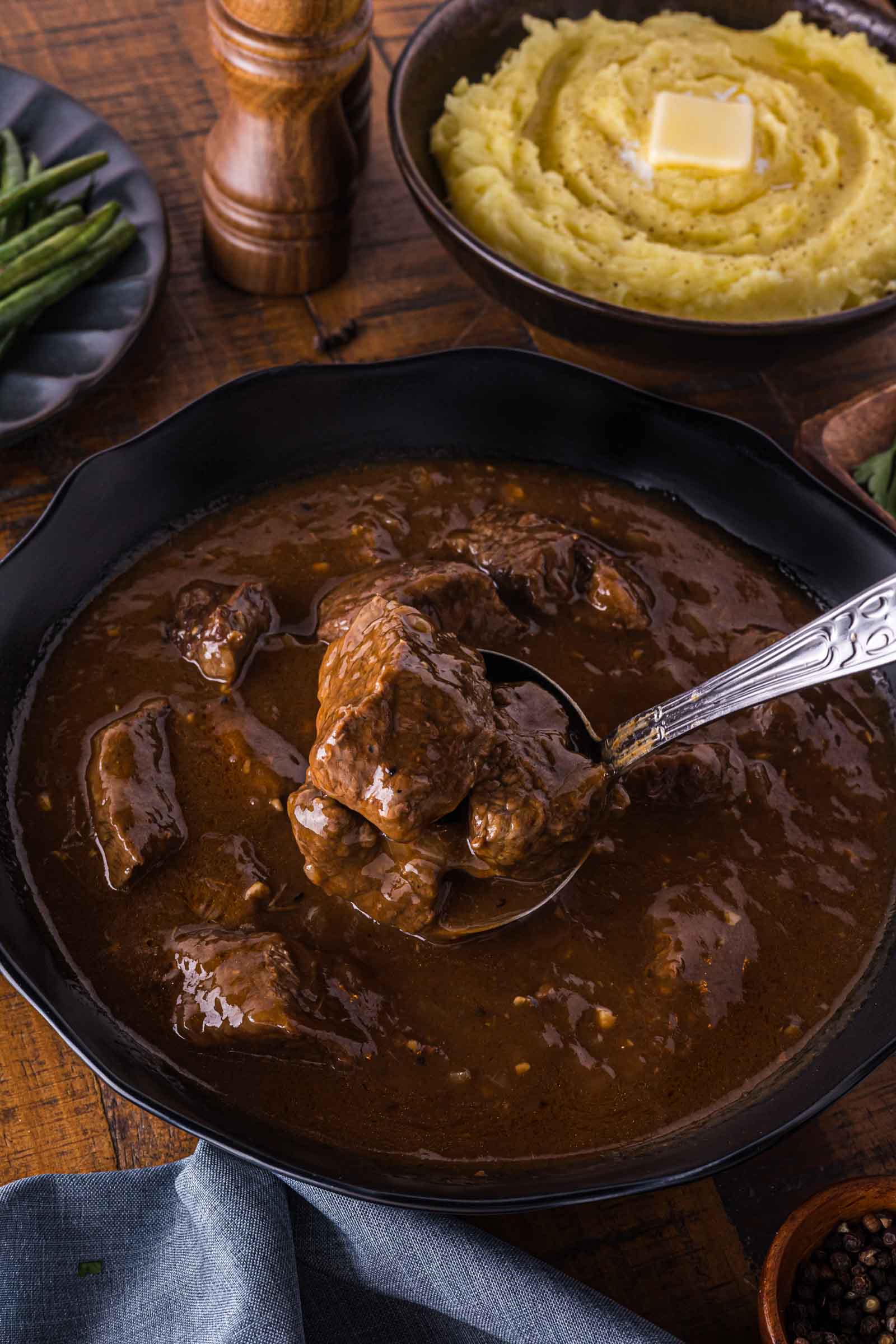 Bowl of beef tips in brown gravy, with a spoon lifting a portion; mashed potatoes with butter and green beans are in the background.