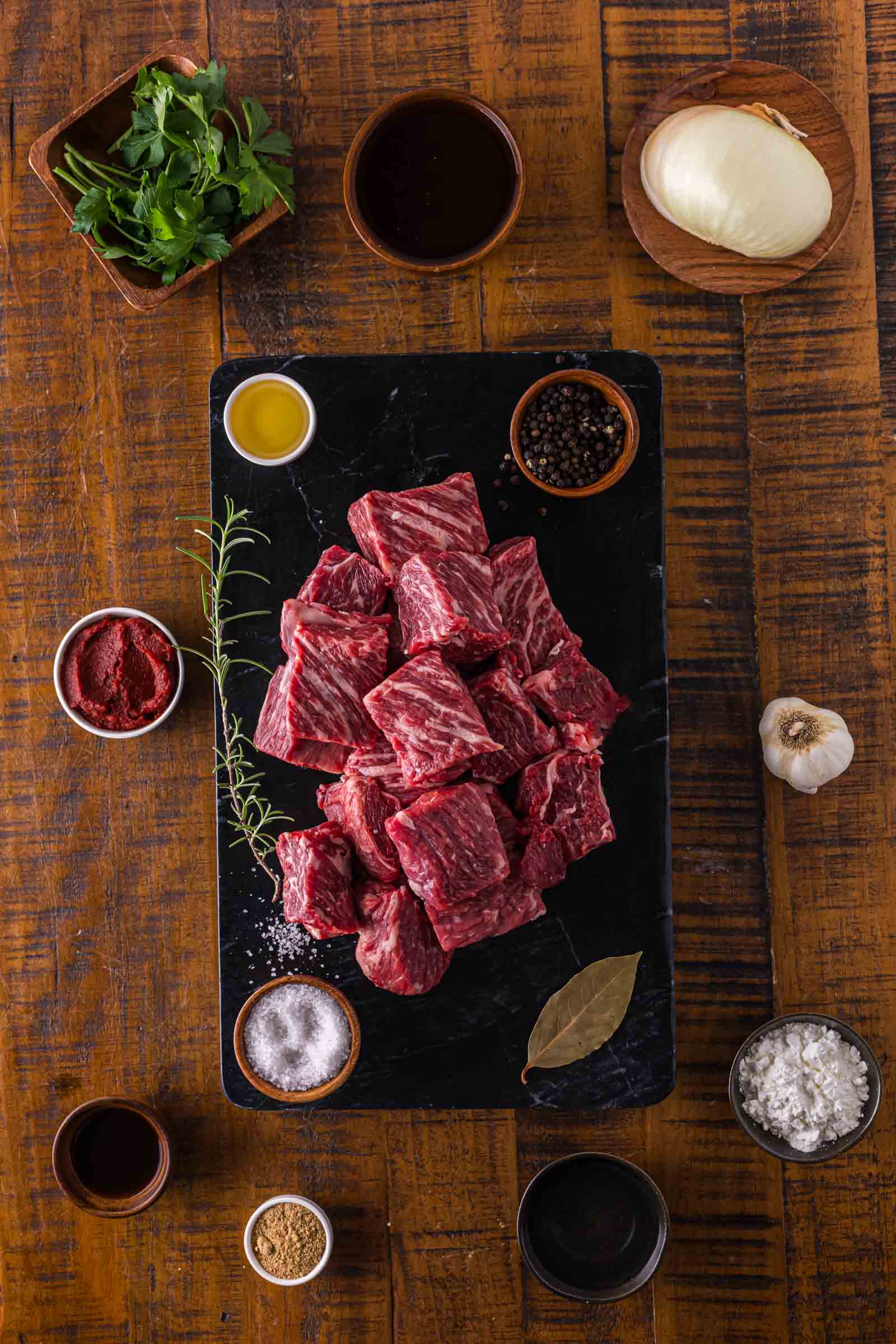 Raw beef chuck pieces on a black slate surrounded by various ingredients including onion, parsley, garlic, tomato paste, bay leaf, spices, and liquids on a wooden table.