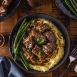A bowl of mashed potatoes topped with beef tip in gravy, garnished with parsley, and served with green beans; additional sides and utensils are on a wooden table.