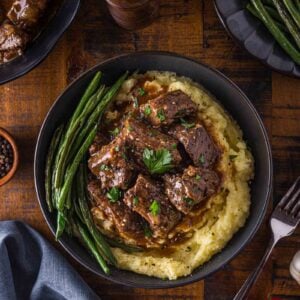 A bowl of mashed potatoes topped with beef tip in gravy, garnished with parsley, and served with green beans; additional sides and utensils are on a wooden table.