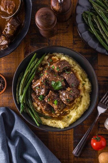 A bowl of mashed potatoes topped with beef tip in gravy, garnished with parsley, and served with green beans; additional sides and utensils are on a wooden table.