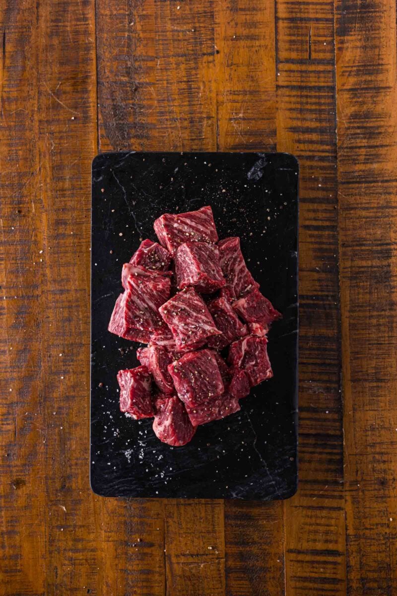 Raw beef chuck seasoned with salt and pepper arranged on a black rectangular tray, set on a wooden surface.