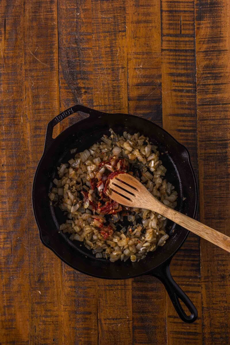 Chopped onions and tomato paste being sautéed in a black cast-iron skillet with a wooden spoon on a wooden surface.