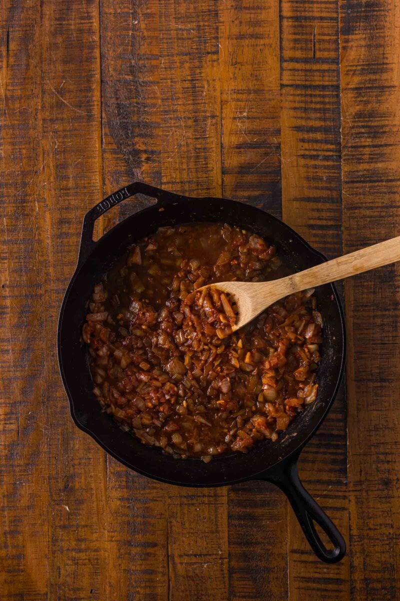 A cast iron skillet with sautéed onions and tomatoes being stirred with a wooden spoon, placed on a rustic wooden surface.