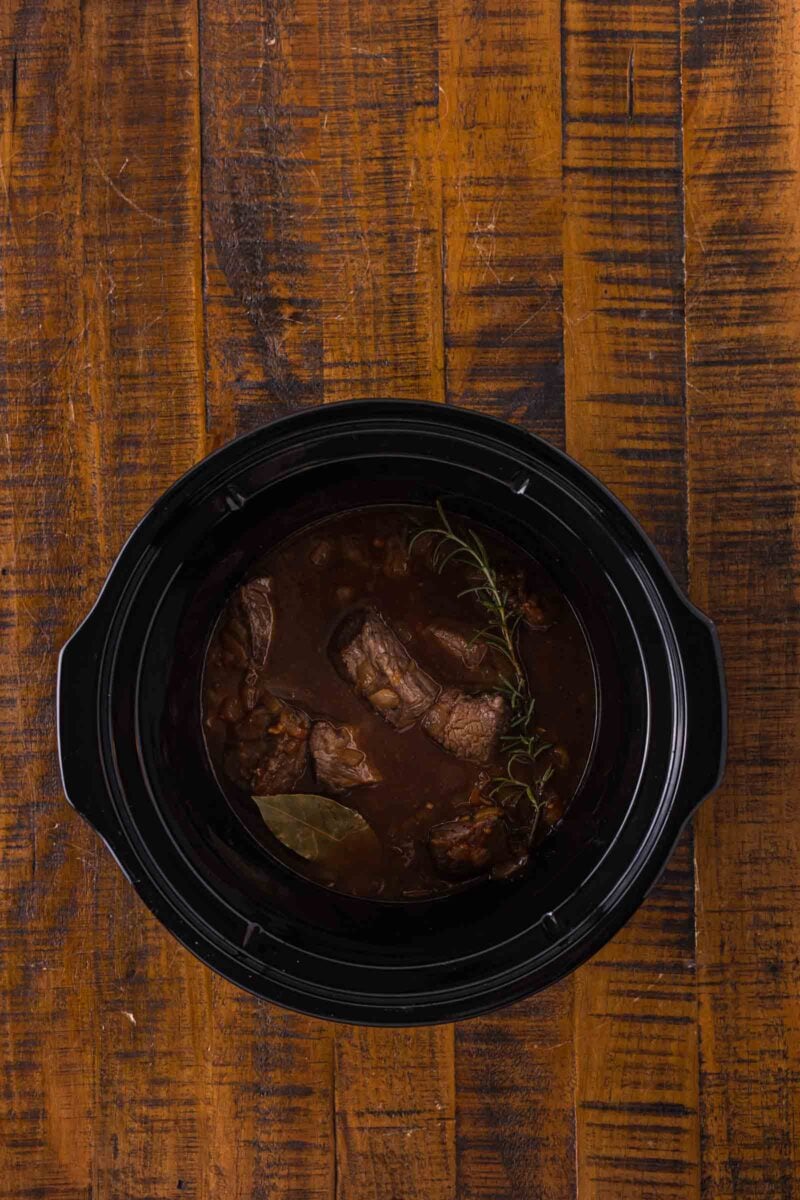 Overhead view of beef tips with herbs in a black slow cooker on a wooden table.
