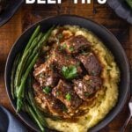 A bowl of mashed potatoes topped with slow cooker beef tips and gravy, served with green beans, on a wooden table. Text reads, "How to make slow cooker beef tips.