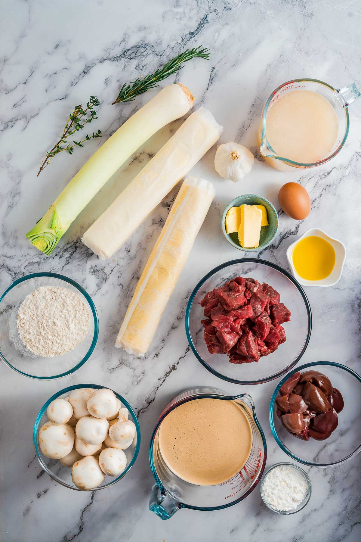 Various raw ingredients arranged on a marble surface, including leeks, pastry, herbs, garlic, butter, egg, flour, oil, beef, lamb kidneys, mushrooms, broth, and cream.
