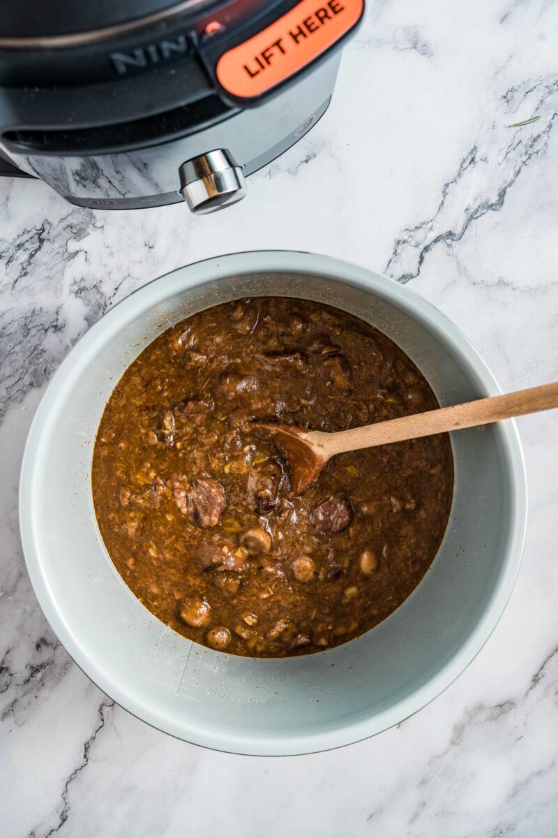 A bowl of guinness meat pie filling with chunks of meat, stirred with a wooden spoon, sits on a marble countertop near a pressure cooker.
