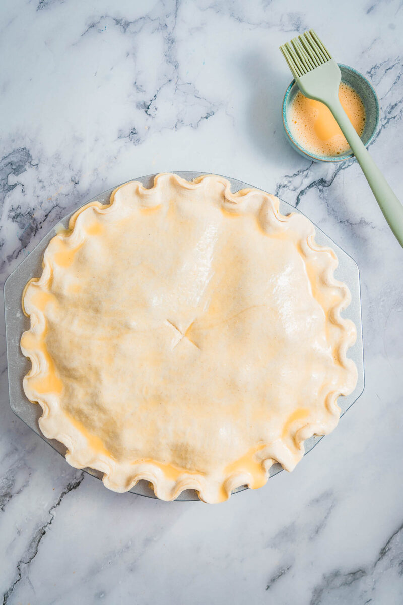 Unbaked steak and kidney pie with crimped edges on a marble surface, next to a small bowl of egg wash and a pastry brush.