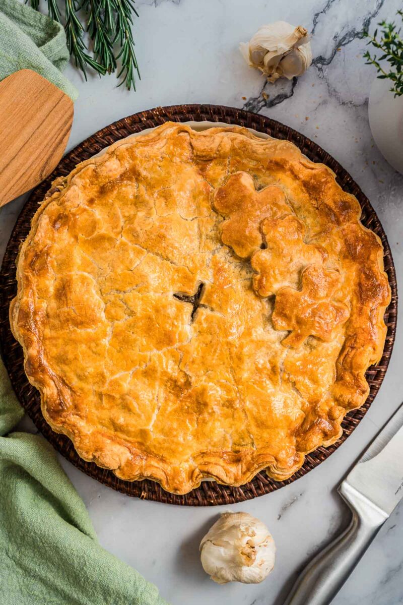 A golden-brown steak and kidney pie with a flaky crust sits on a round tray, surrounded by a knife, garlic bulbs, a green cloth, and sprigs of herbs on a marble surface.