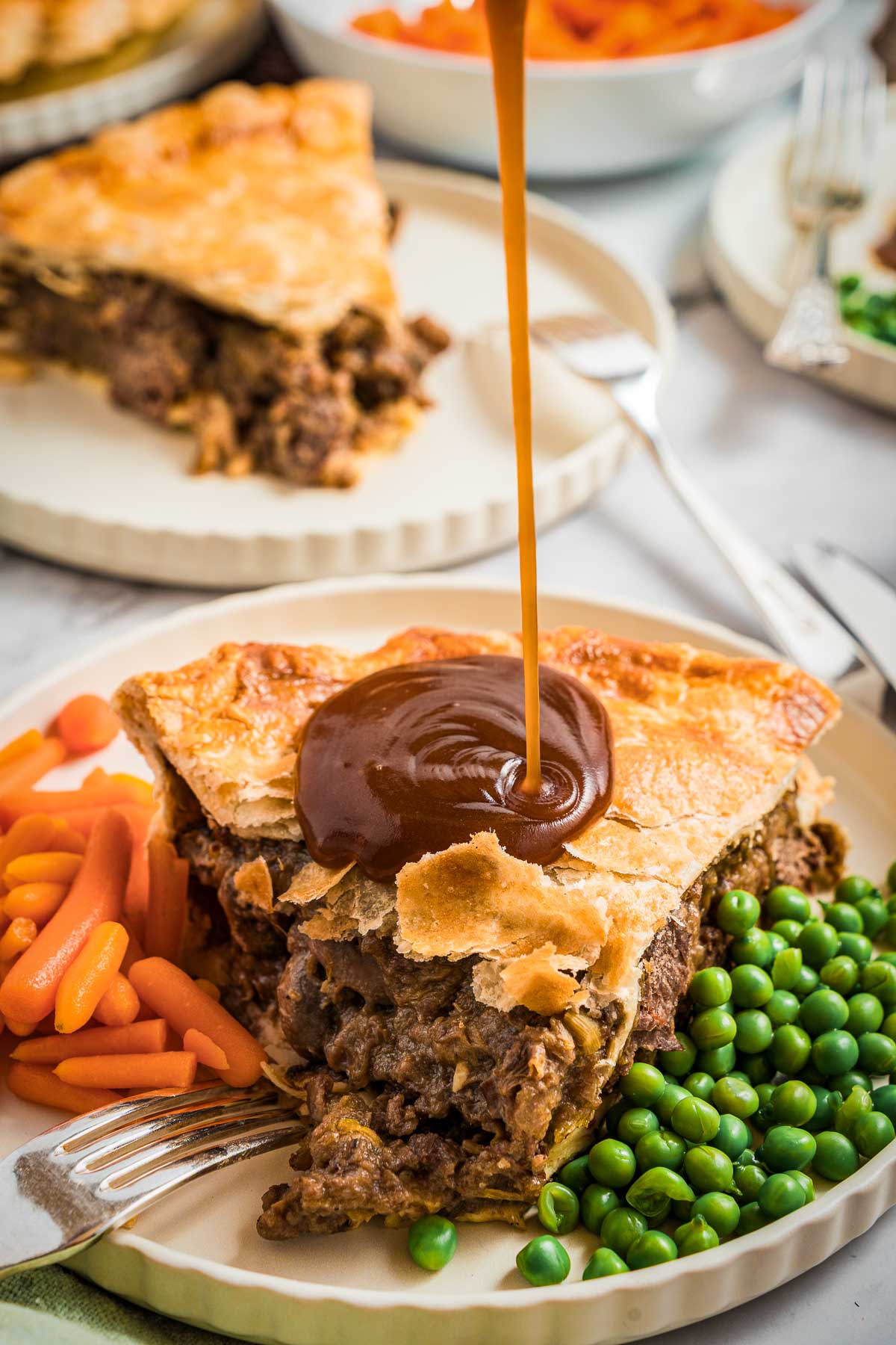 A slice of steak and kidney pie with golden crust is served with peas and carrots, while brown gravy is being poured on top.