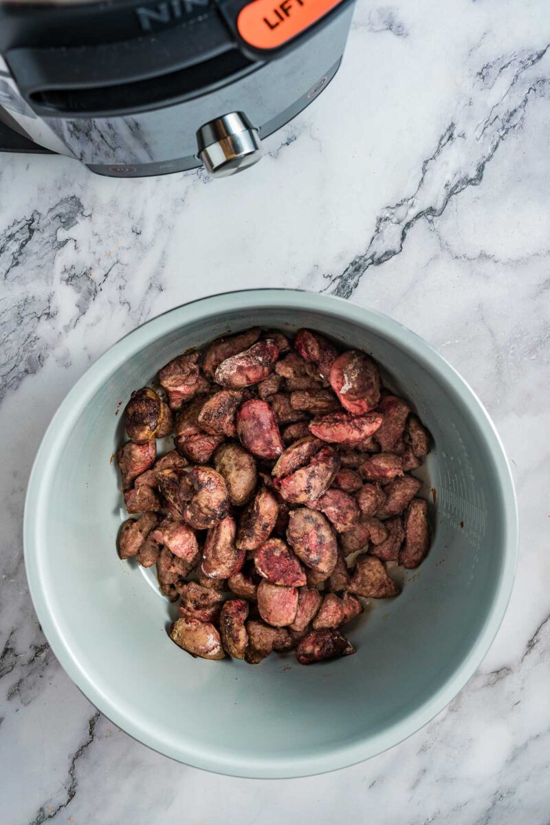 A white bowl containing frozen kidneys and steak pieces on a marble countertop, with part of an air fryer visible in the upper left corner.
