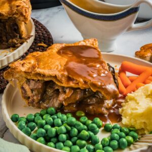 A plate with a slice of steak and kidney pie topped with gravy, mashed potatoes, green peas, and baby carrots; gravy boat and another plate in the background.