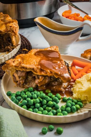 A plate with a slice of steak and kidney pie topped with gravy, mashed potatoes, green peas, and baby carrots; gravy boat and another plate in the background.