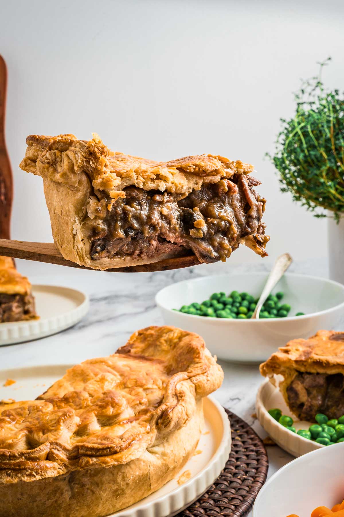 A slice of steak and kidney pie being served from a larger pie, with bowls of peas and carrots in the background on a marble table.