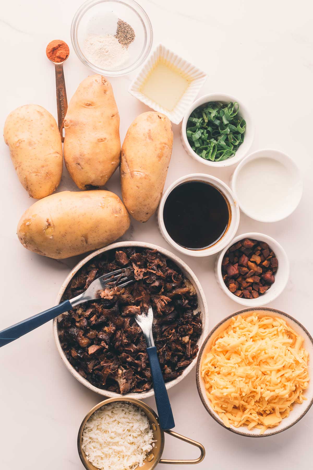 Overhead view of ingredients on a white surface, including potatoes, shredded cheese, cooked meat, green onions, butter, seasonings, and small bowls of sauces and cream.