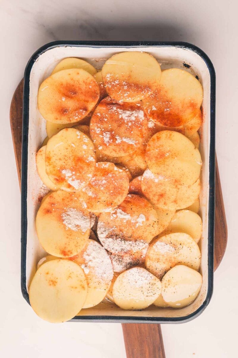 A baking dish filled with sliced potatoes topped with flour, paprika, and black pepper, placed on a wooden board.