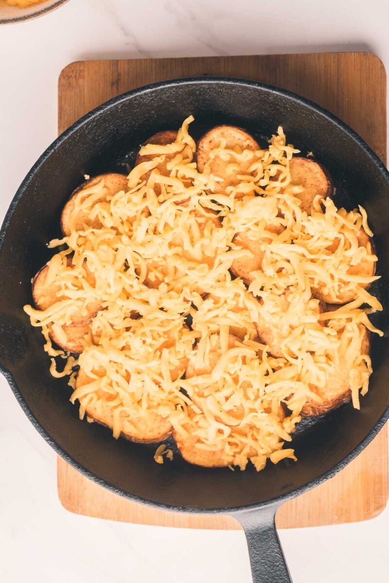 A cast iron skillet on a wooden board filled with sliced potatoes topped with shredded cheese.