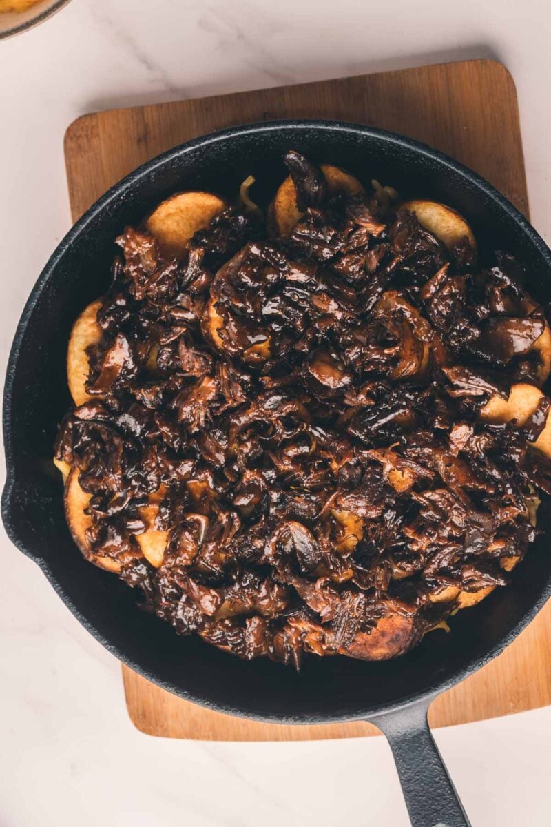 A cast iron skillet filled with sliced potatoes topped with a dark, rich mushroom and beef mixture, placed on a wooden board.