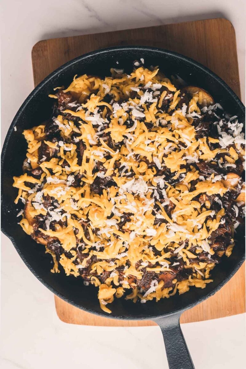 A cast iron skillet filled with shredded cheese and caramelized onions sits on a wooden board over a marble surface.