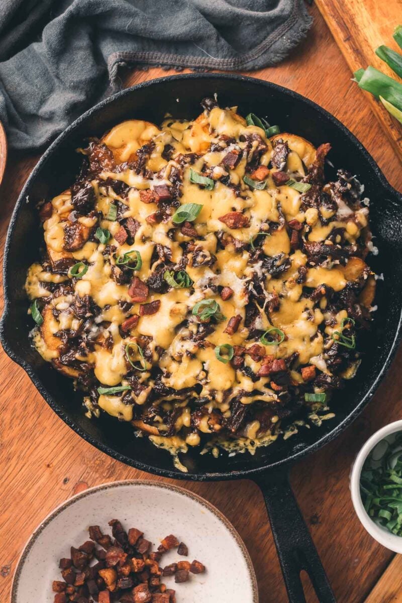 A cast iron skillet filled with cheesy potatoes, sautéed mushrooms, bacon bits, and green herbs, placed on a wooden table.