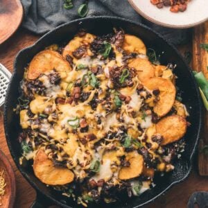 A cast iron skillet filled with sliced potatoes, melted cheese, diced vegetables, and green onions, with extra toppings in small bowls nearby on a wooden table.