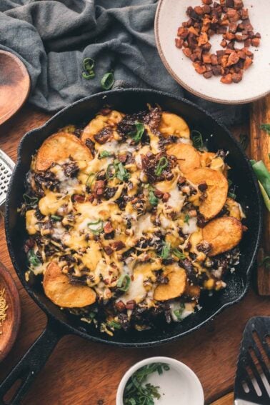 A cast iron skillet filled with sliced potatoes, melted cheese, diced vegetables, and green onions, with extra toppings in small bowls nearby on a wooden table.