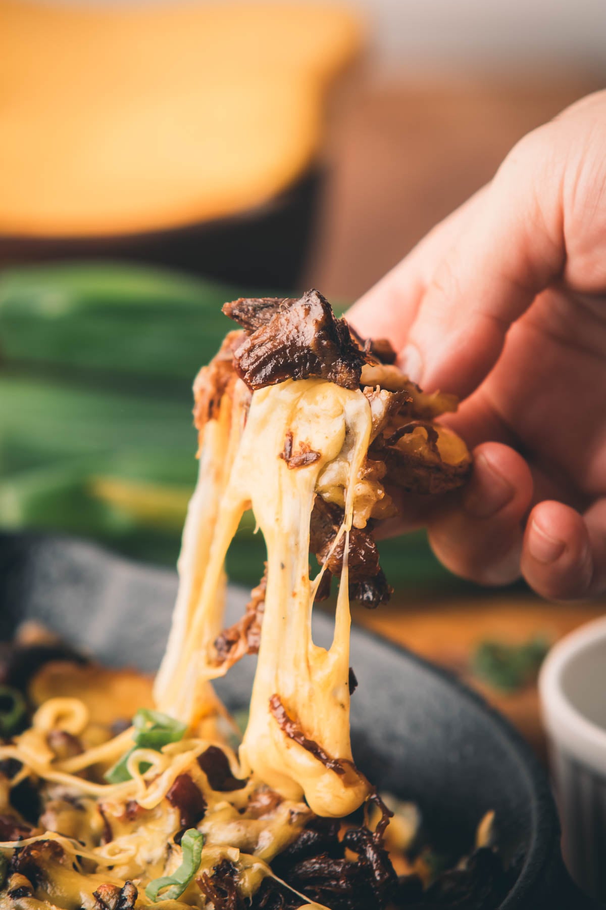 A hand lifts a serving of cheesy, melted beef and noodles from a bowl, with cheese stretching between the food and the bowl.