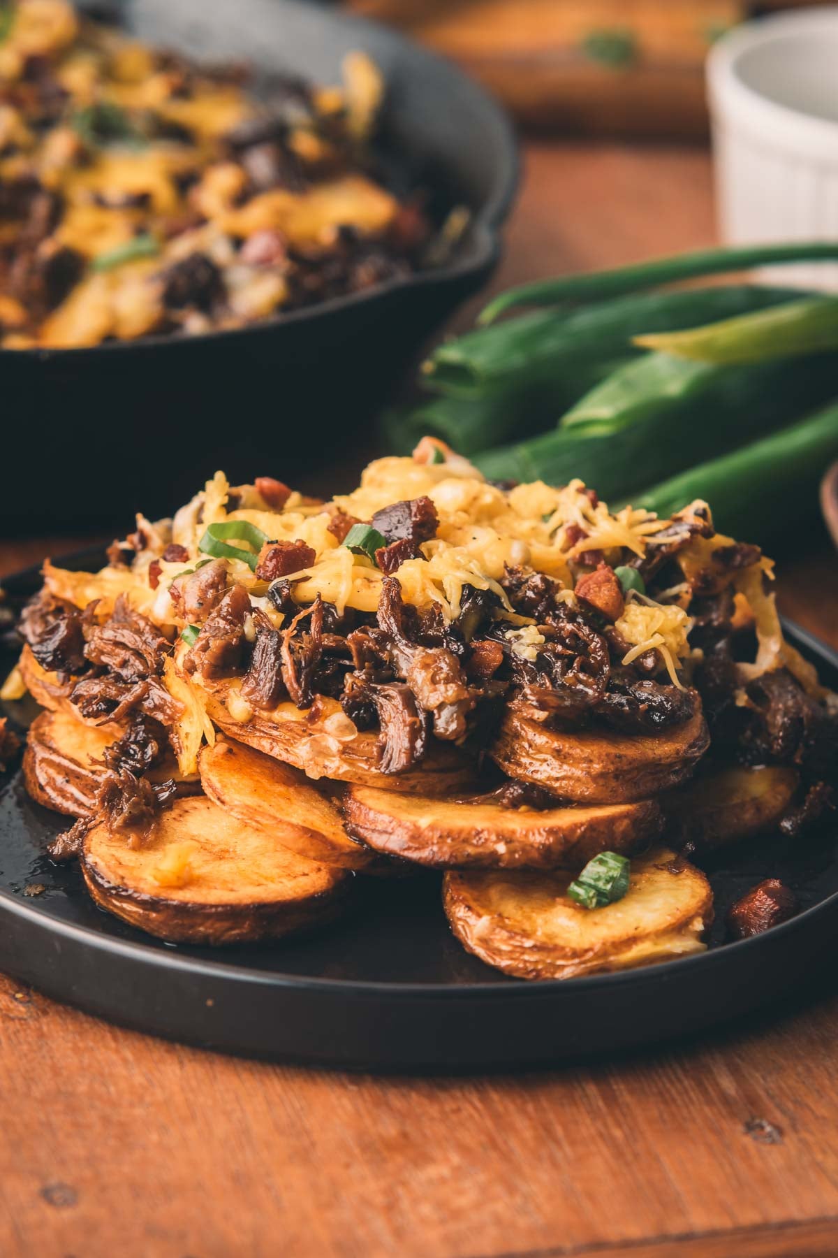 A plate of crispy sliced potatoes topped with shredded cheese, chopped green onions, and cooked meat sits on a wooden table.