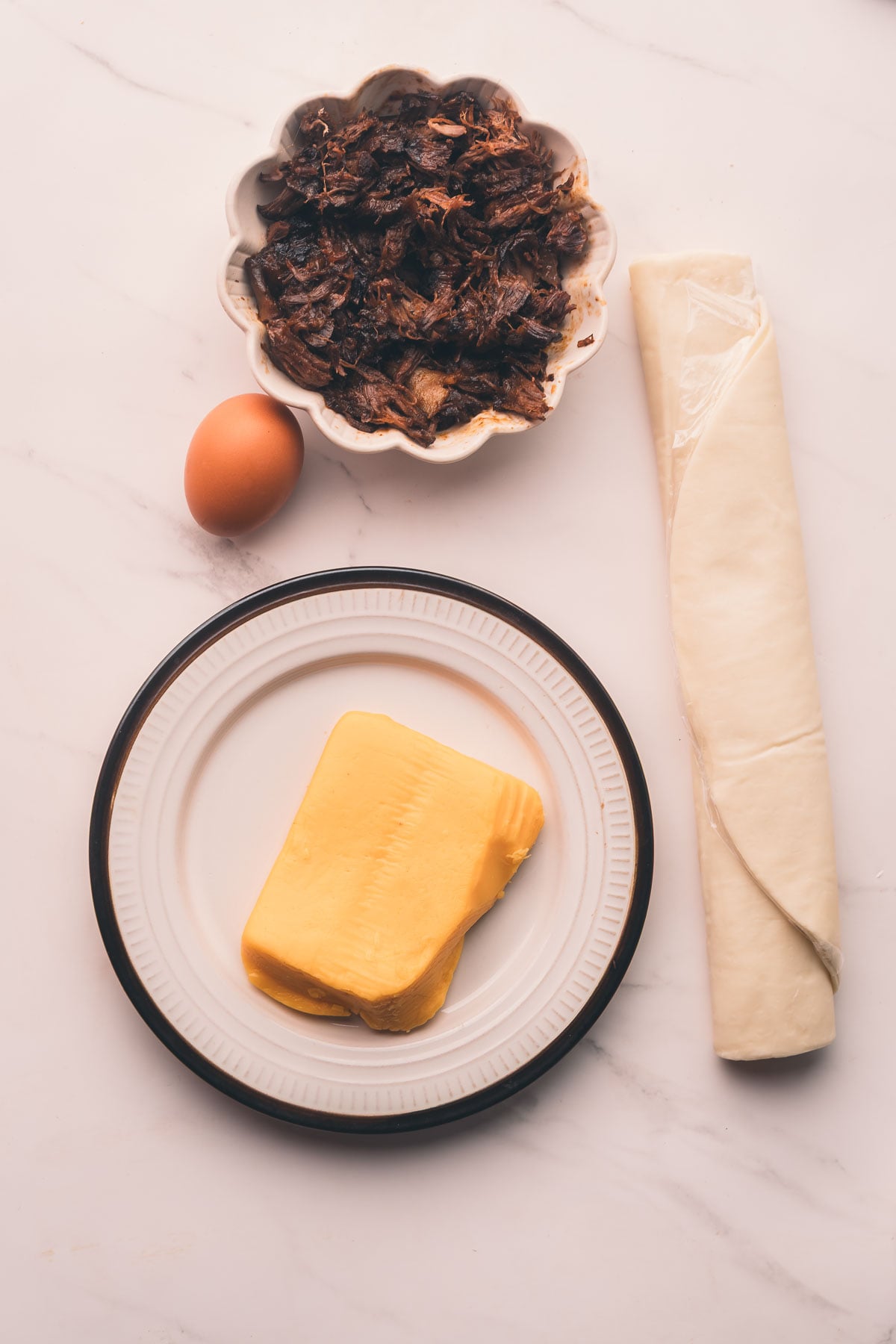 A plate with cheese, a raw egg, a bowl of shredded braised beef shanks, and a roll of puff pastry on a white marble surface.