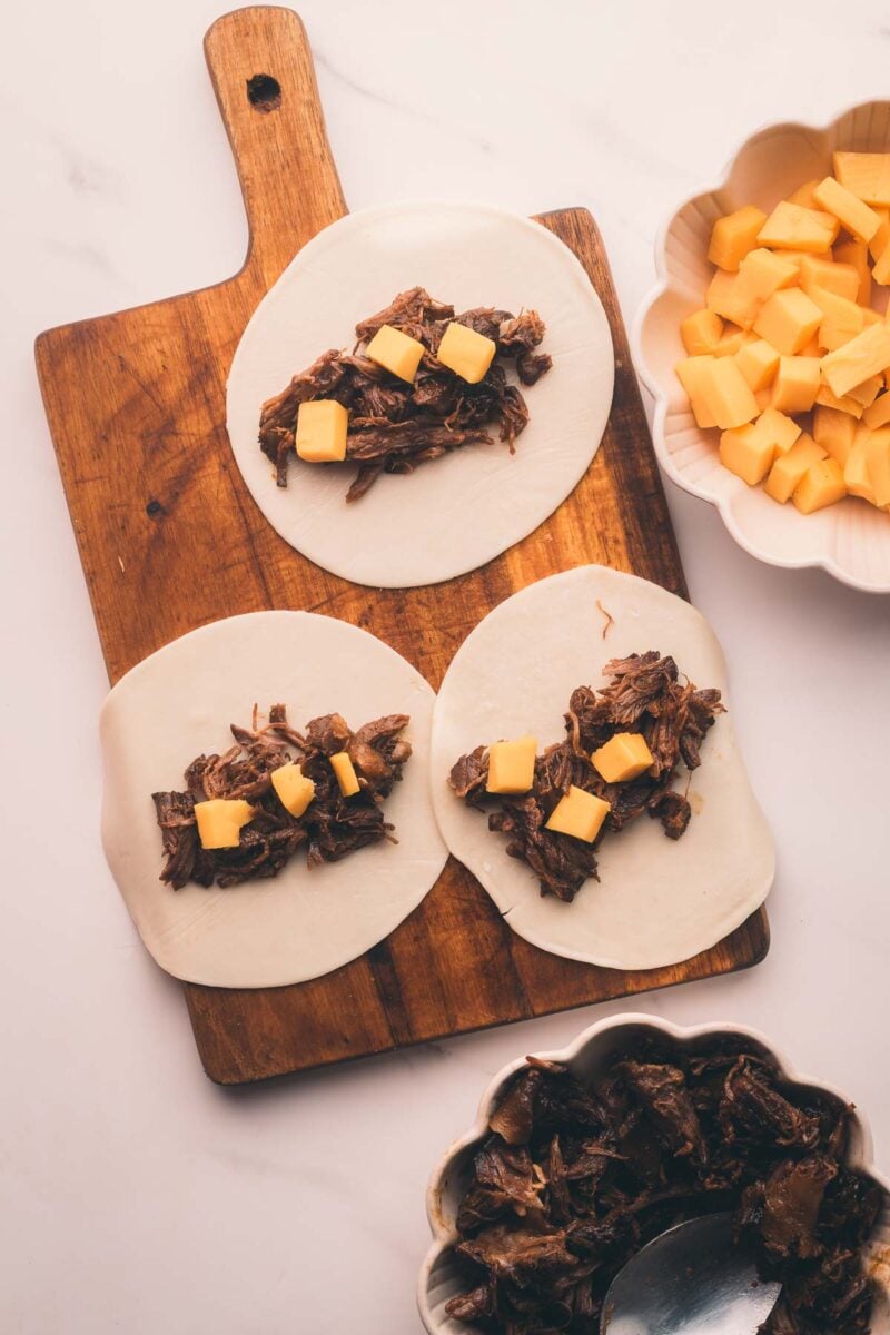 Three uncooked dough circles topped with stout braised beef and small cheese cubes on a wooden board, with bowls of more shredded meat and diced cheese nearby.