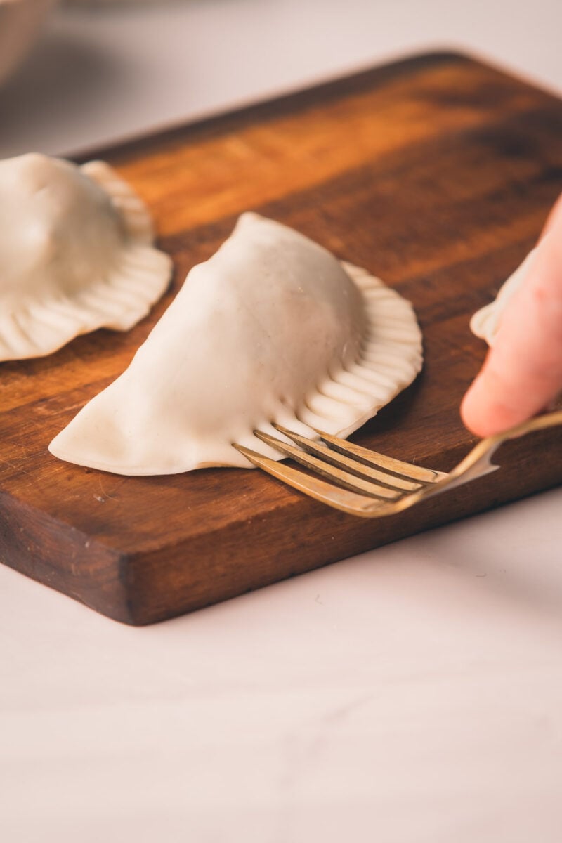 A hand presses the edge of a hand pie with a fork on a wooden board, sealing the dough. Another uncooked dumpling is visible in the background.