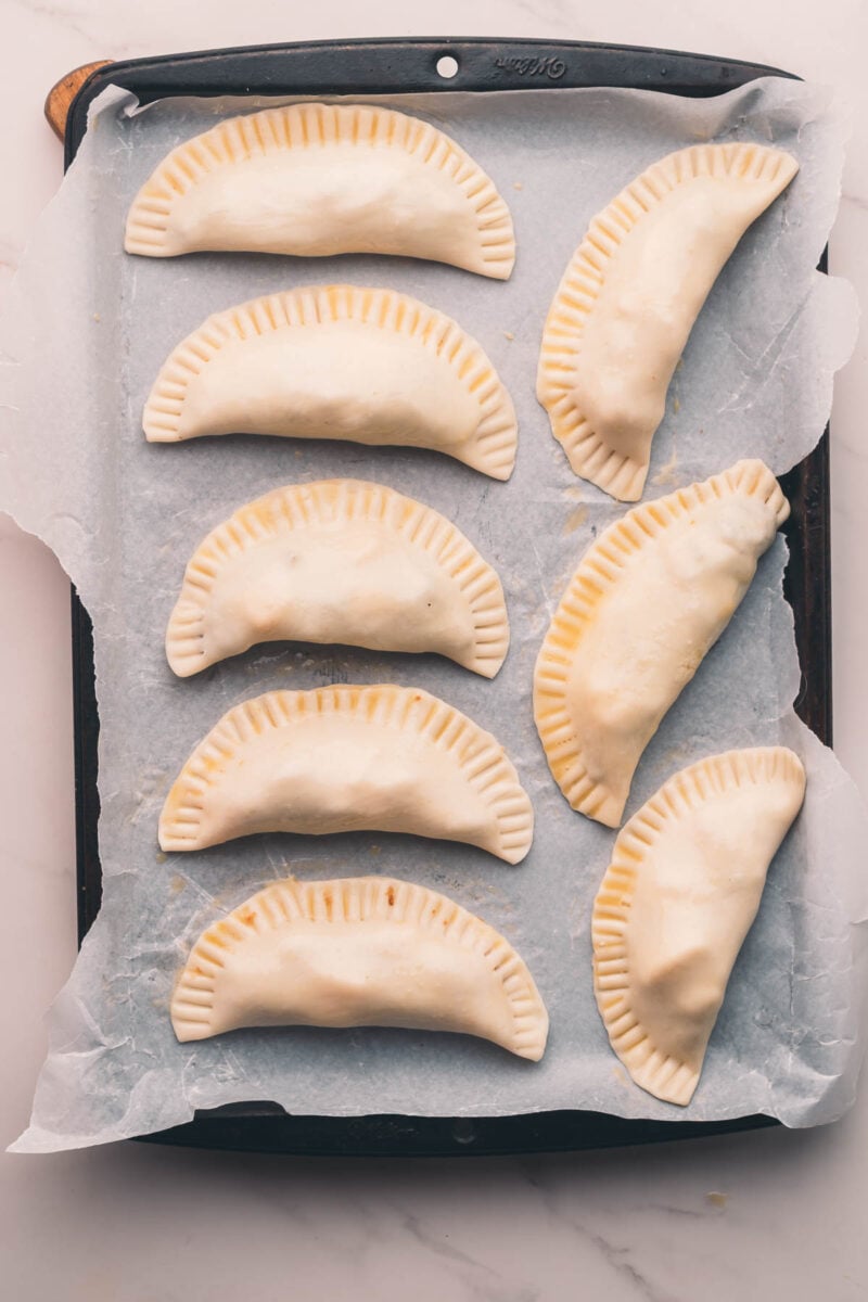 Eight uncooked, crimped hand pies or hand pies are arranged on a parchment-lined baking sheet.