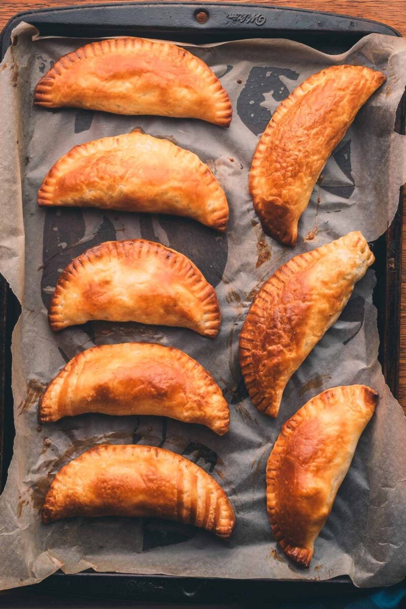 A baking tray lined with parchment paper holds seven golden-brown baked hand pies arranged in rows.