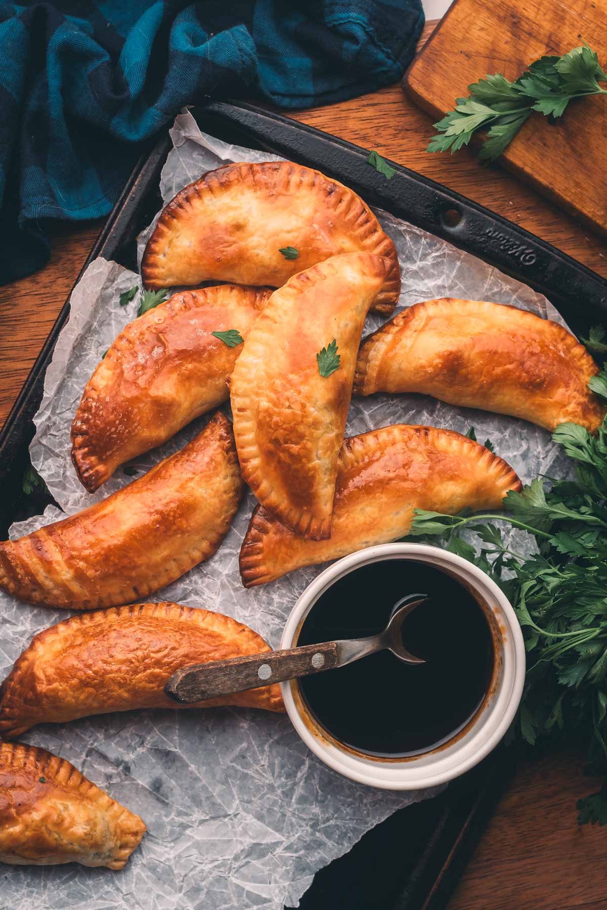 Golden-brown braised beef baked hand pies are arranged on a parchment-lined tray with a bowl of dark dipping sauce and a garnish of fresh parsley.