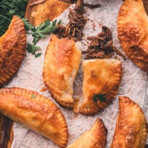 Five baked hand pies arranged on parchment paper, one split open to show shredded meat filling, with fresh parsley and a bowl of dark dipping sauce nearby.