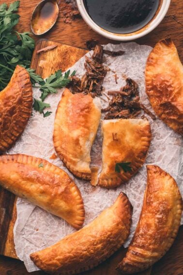 Five baked hand pies arranged on parchment paper, one split open to show shredded meat filling, with fresh parsley and a bowl of dark dipping sauce nearby.