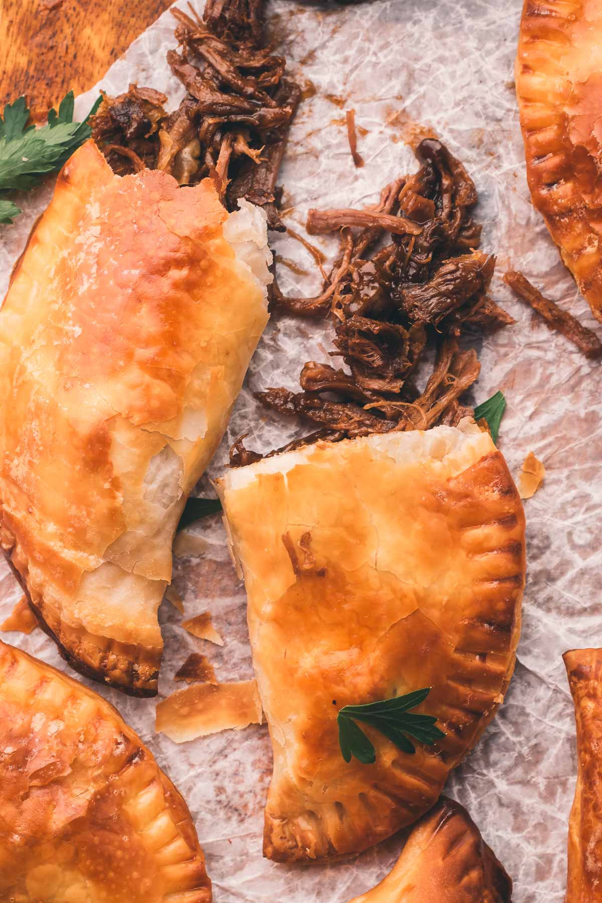 Golden brown stout beef hand pies are shown on parchment paper, one is broken open to reveal shredded beef filling. Parsley garnish is visible.