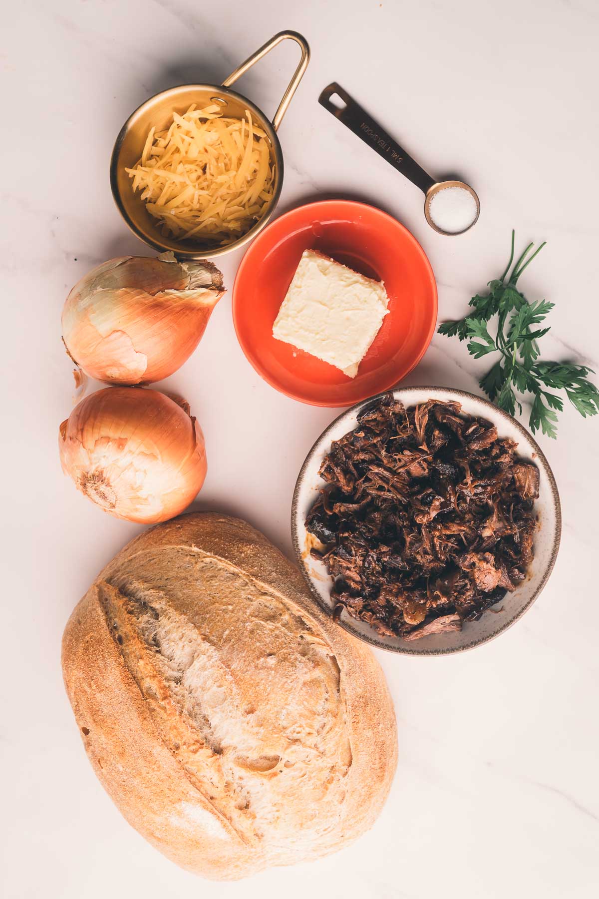 A loaf of bread, two onions, shredded cheese in a measuring cup, a square of butter on a small plate, parsley, and a bowl of shredded meat on a white surface.