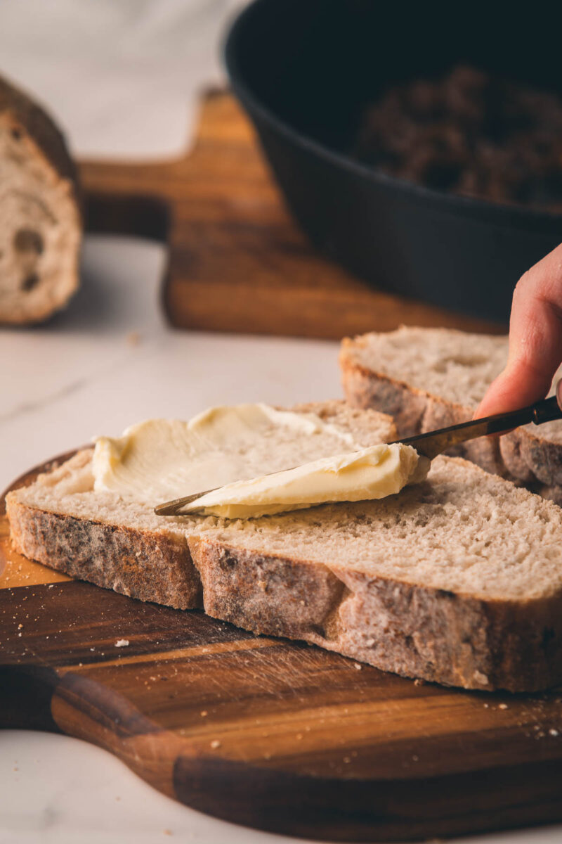A hand spreads butter on a slice of rustic bread with a knife, with more bread and a black pan in the background on a wooden board.