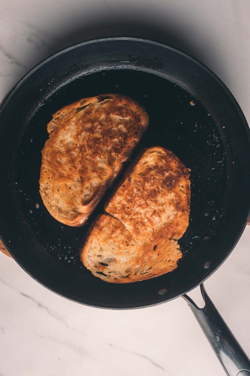Two grilled cheese sandwiches being cooked in a black frying pan on a white marble countertop.