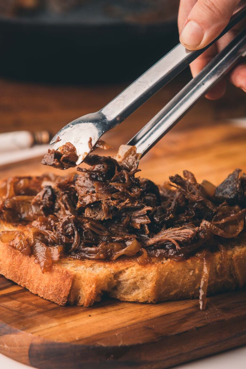 A hand uses tongs to place shredded beef on a slice of toasted bread on a wooden cutting board.