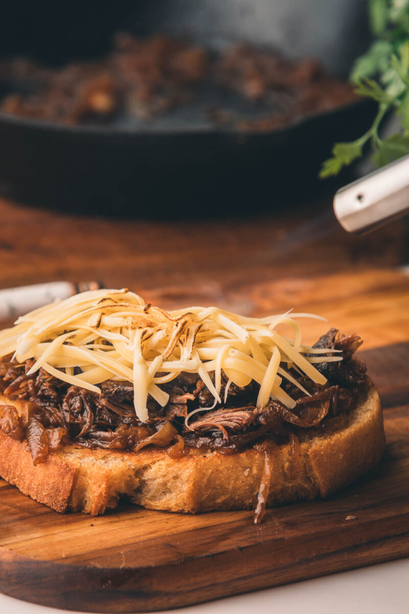 An open-faced sandwich with shredded beef and a generous topping of grated cheese on toasted bread, placed on a wooden cutting board.