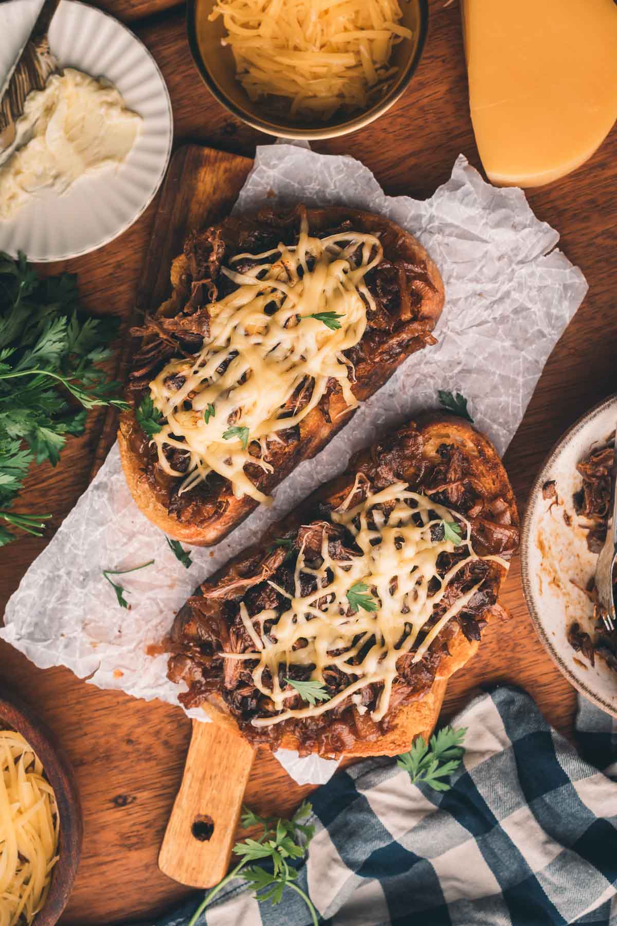 Two open faced sandwiches topped with shredded meat, melted cheese, and fresh herbs are placed on parchment paper, surrounded by bowls of grated cheese and fresh parsley.