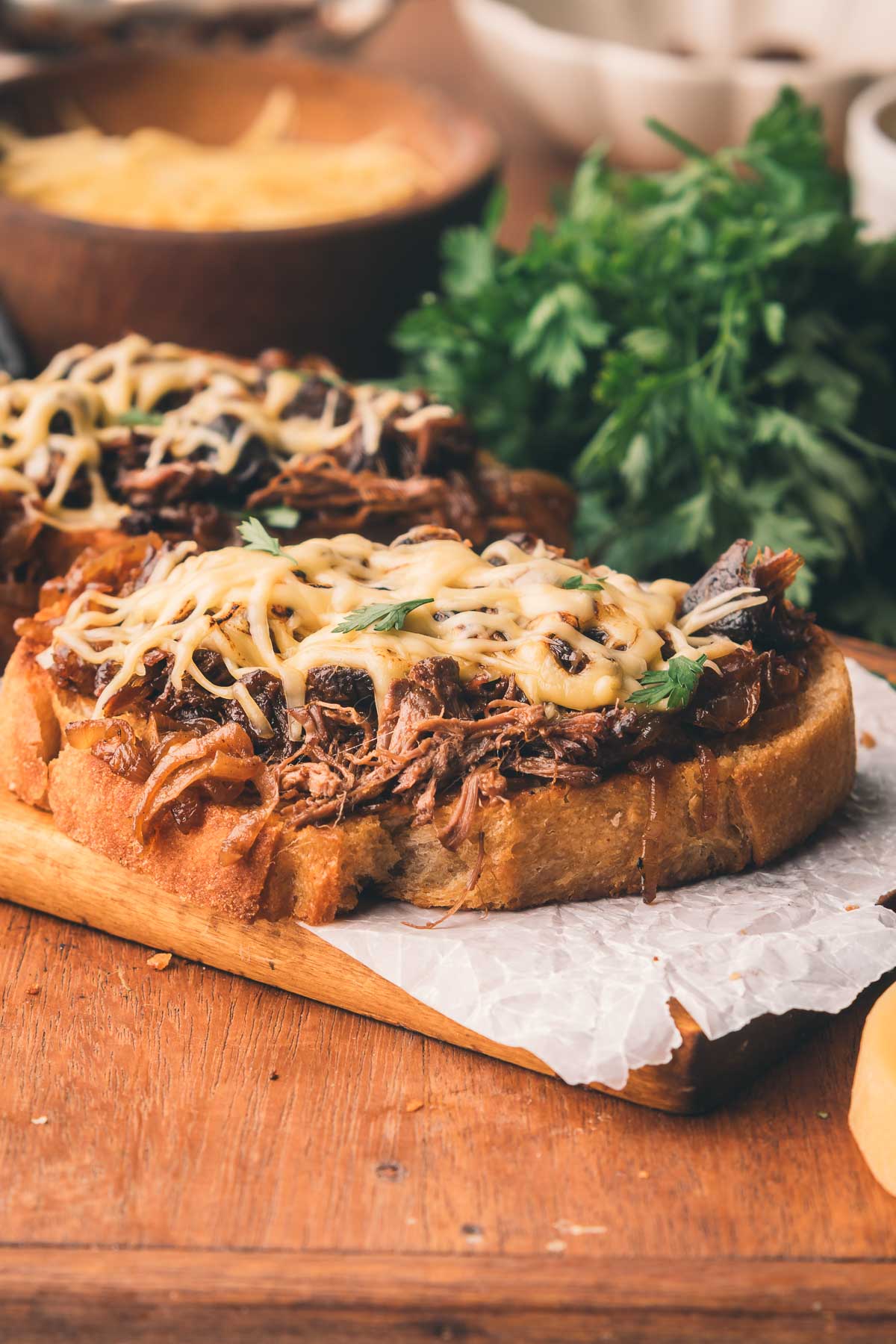 An open faced sandwich topped with shredded braised beef, sautรฉed onions, and melted cheese on a wooden board, with parsley in the background.
