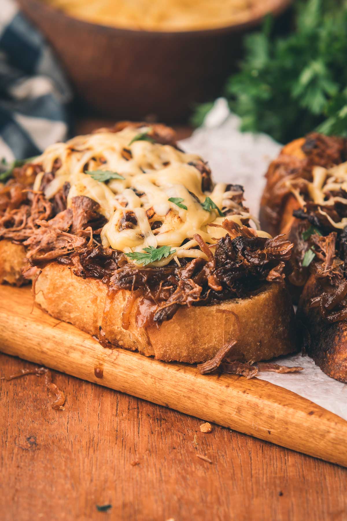 An open faced braised beef sandwich with melted cheese, and chopped herbs on a wooden cutting board.
