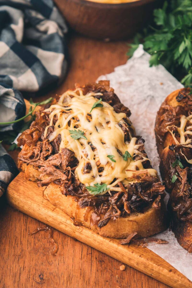 Open-faced sandwich topped with shredded meat, melted cheese, and herbs on a wooden board, with a checkered cloth and greens in the background.