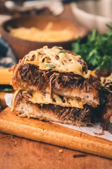 Close-up of an oped faced sandwich stacked with shredded beef, melted cheese, and caramelized onions on toasted bread, served on a wooden board.