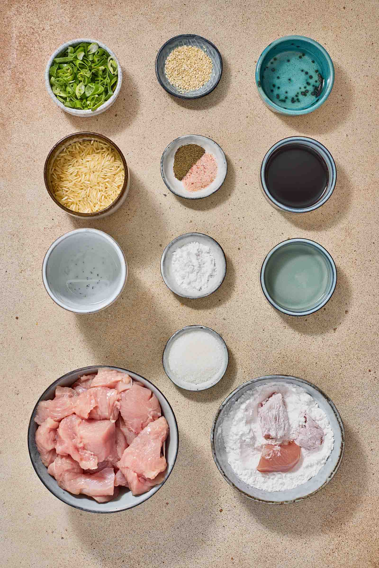 Top-down view of bowls containing sliced green onions, sesame seeds, rice, seasonings, soy sauce, water, chicken pieces, cornstarch, and other ingredients arranged on a textured surface.